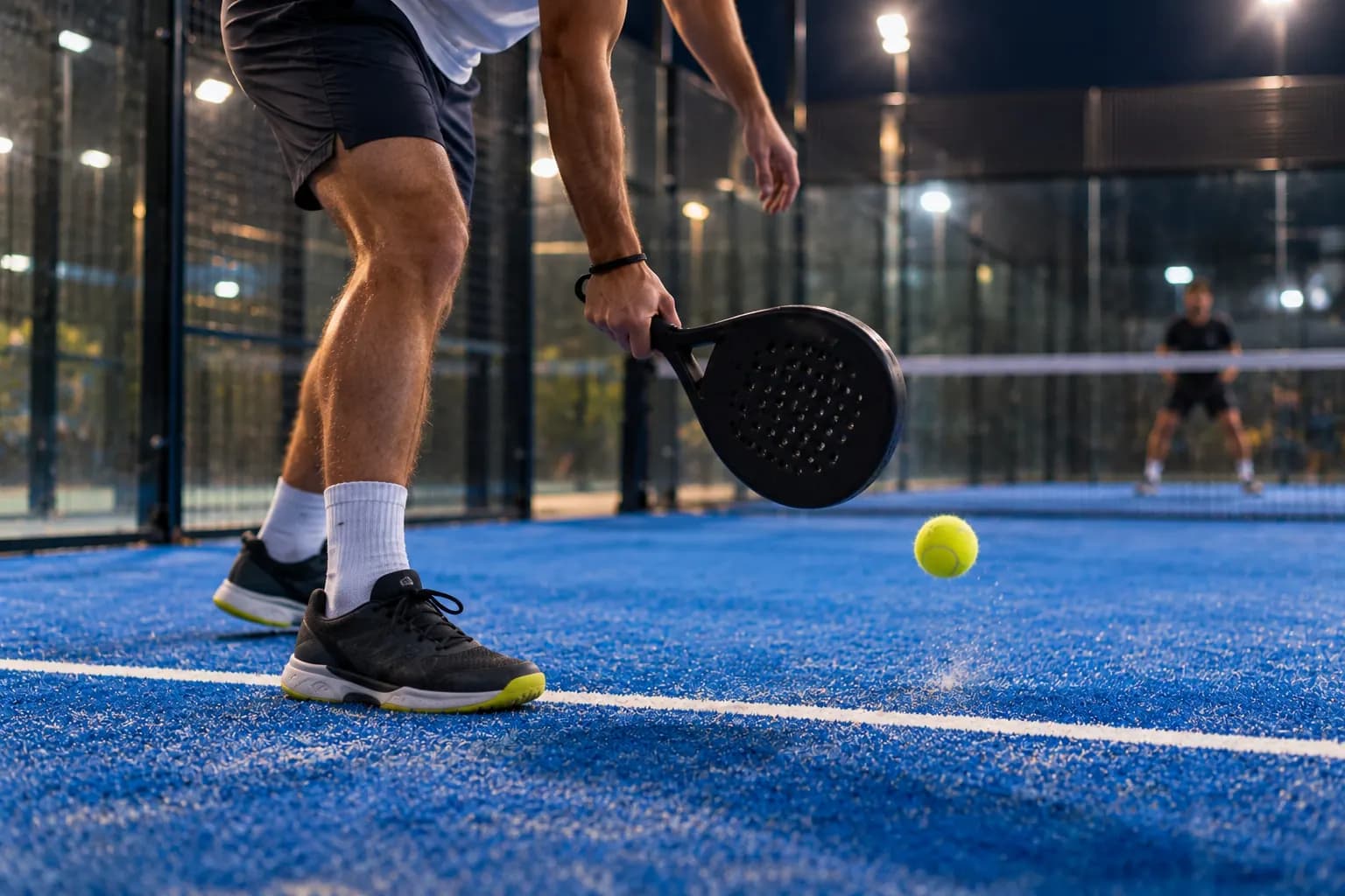 Nahaufnahme von Beinen und schwarzem Schläger eines Padelspielers, während der gelbe Ball unter Flutlicht auf dem blauen Kunstrasen aufspringt; auf der anderen Netzseite wartet ein Gegner.
